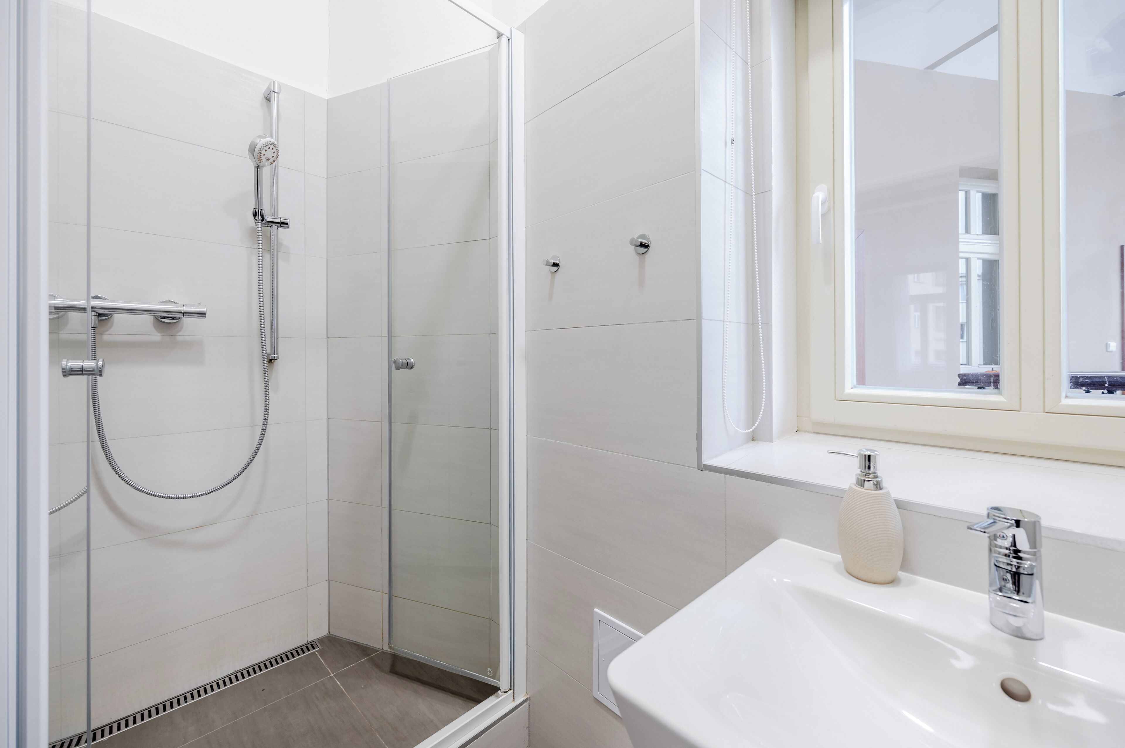 Ein modernes Badezimmer mit einer gläsernen Duschkabine, einem weißen Waschbecken und einem Fenster darüber.
