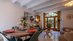 Un comedor con una mesa redonda, sillas y un jarrón de flores. Hay un sofá y una mesita con una planta. Se ve una ventana grande con cortinas.