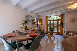 Ein Essbereich mit rundem Tisch, Stühlen und einem Blumenstrauß. Ein Sofa und ein kleiner Tisch mit einer Pflanze sind ebenfalls zu sehen. Ein großes Fenster mit Vorhängen ist erkennbar.