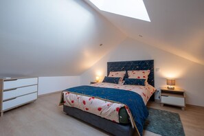 A bedroom with a sloped ceiling, a bed with a floral bedspread, two bedside tables with lamps, and a dresser. - Gîte La Sagnette 4/6 persons (LANUEJOLS)
