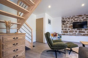 A modern living room with a wooden staircase, a stone wall, a flat-screen TV, and a green upholstered chair. - Gîte La Sagnette 4/6 persons (LANUEJOLS)