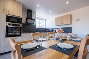 A modern kitchen with wooden cabinets, stainless steel appliances, and a dining area with a wooden table set for six. - Gîte La Sagnette 4/6 persons (LANUEJOLS)