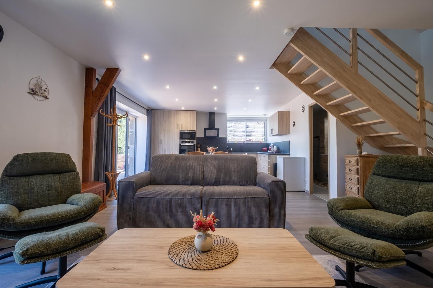 A modern living room with a sofa, two armchairs, and a coffee table. A kitchen is visible in the background with a staircase leading to an upper level.