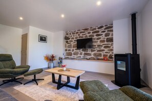 A living room with a stone wall, a TV mounted above it, a black wood-burning stove, and two green armchairs. - Gîte La Sagnette 4/6 persons (LANUEJOLS)