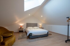 A bedroom with a sloped ceiling, a bed with a grey headboard, two bedside lamps, a wooden nightstand, a sofa, and a floor lamp. - La Sagnette Vacation Rental for 4–6 People (LANUEJOLS)