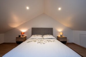 A bedroom with a sloped ceiling, a bed with a leaf-patterned bedspread, two bedside tables with lamps, and a wooden floor. - Gîte La Sagnette 4/6 persons (LANUEJOLS)