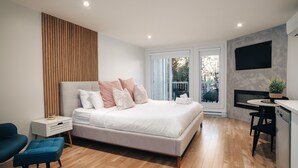 A modern bedroom with a bed, bedside table, chair, and a small dining area with a TV. - LAO LOFT 106 by Gestion ELITE (Mont-Tremblant)