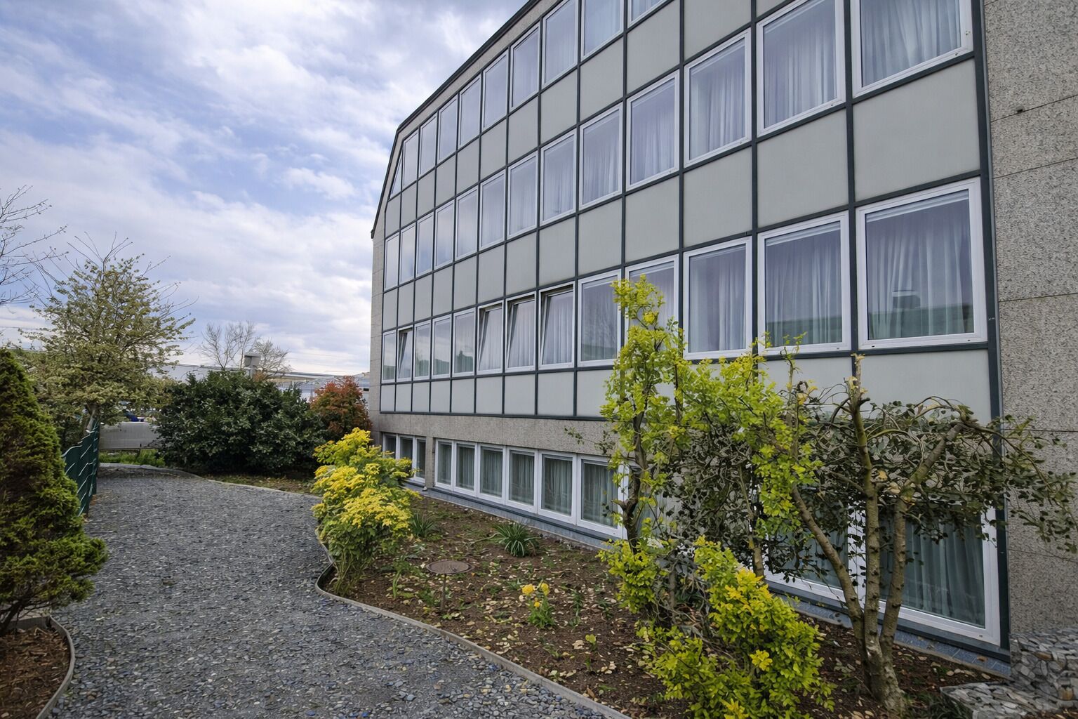 A modern building with a grid of windows, a gravel path, and various shrubs.