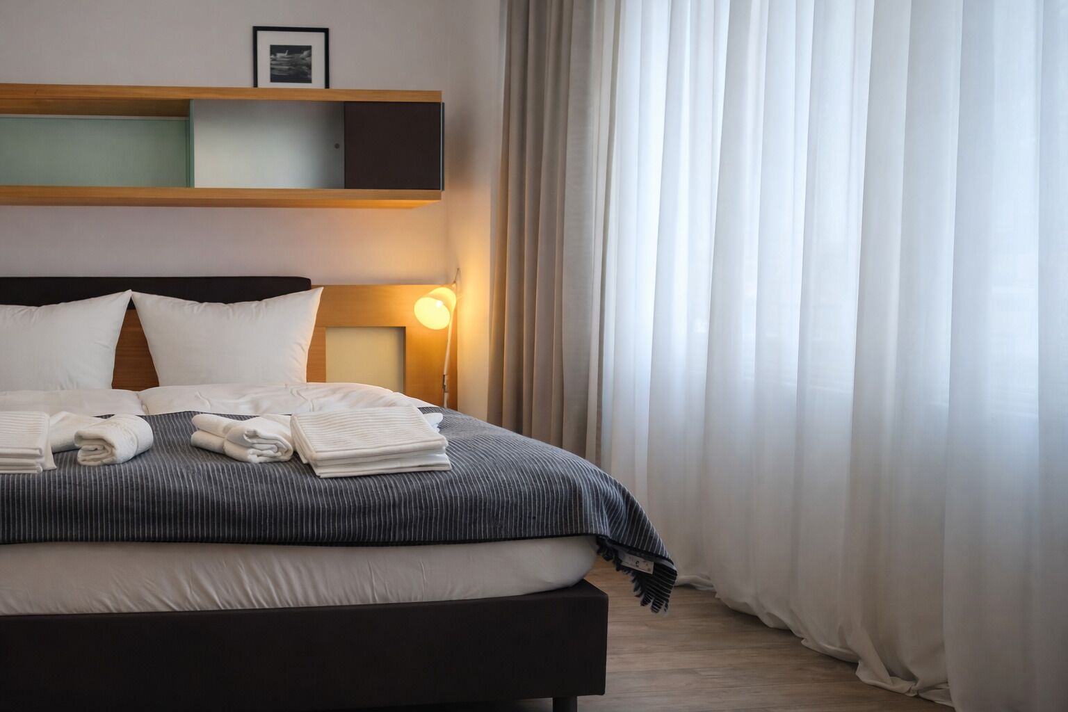 Una cama bien hecha con almohadas y toallas dobladas, una lámpara de noche y un cuadro enmarcado en un estante sobre la cama.