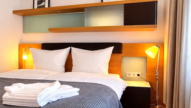 A hotel room with a bed, bedside lamps, a flat-screen TV, and a wall-mounted shelf. - Check-Inn Hotel - Offenbach (Offenbach am Main)