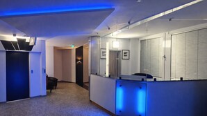 A hotel reception area with a reception desk, a black leather chair, a coat rack, and a blue-lit wall. - Check-Inn Hotel - Offenbach (Offenbach am Main)
