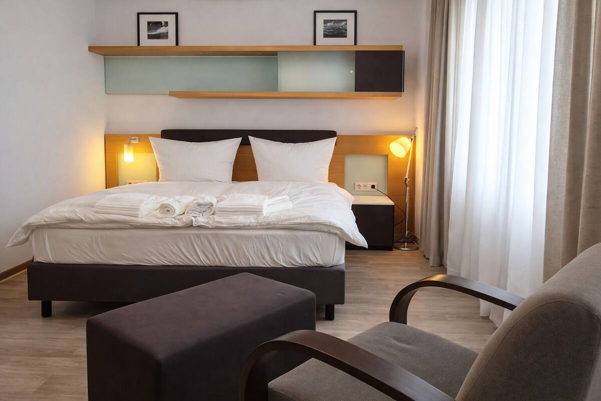 Un lit bien fait, avec du linge de lit blanc, une table de chevet avec une lampe et une chaise dans une chambre d’hôtel moderne.