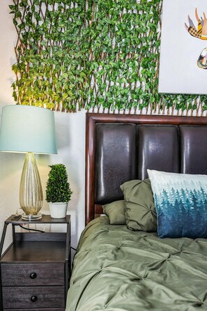 Ein Schlafzimmer mit einem Bett, einem Nachttisch, einer Lampe, einer kleinen Topfpflanze und einer an der Wand befestigten grünen Dekoration mit Blättern.
