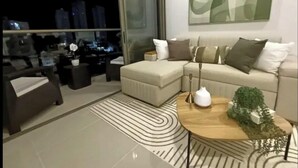 A modern living room with a sectional sofa, a coffee table, and a view of the cityscape through large windows. - Zen Oceanview Apartment Reserva del Mar 2, Santa Marta, Colombia (Gaira)