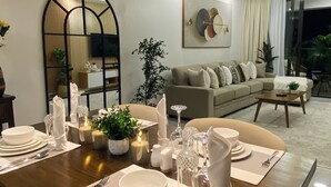 Uma sala de jantar com mesa de madeira posta para quatro pessoas, um sofá ao fundo e um espelho grande na parede.