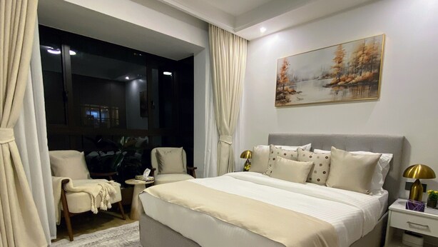 Quarto com cama grande, mesa de cabeceira, cadeira, janela com cortinas e um quadro na parede.