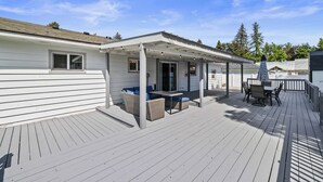 Een overdekt terras buiten met zitplaatsen en een tafel, omgeven door een houten terras en een wit gebouw.