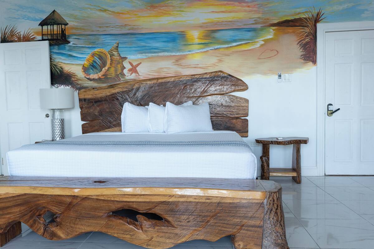 Un dormitorio con cabecera de madera, cama con ropa de cama blanca, una mesita de noche, una lámpara y un mural con escena de playa.