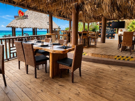 Un’area lounge di un ristorante con tetto di paglia, tavoli in legno, sedie in vimini e vista sull'oceano.. Tiamo Beach Resort and Spa