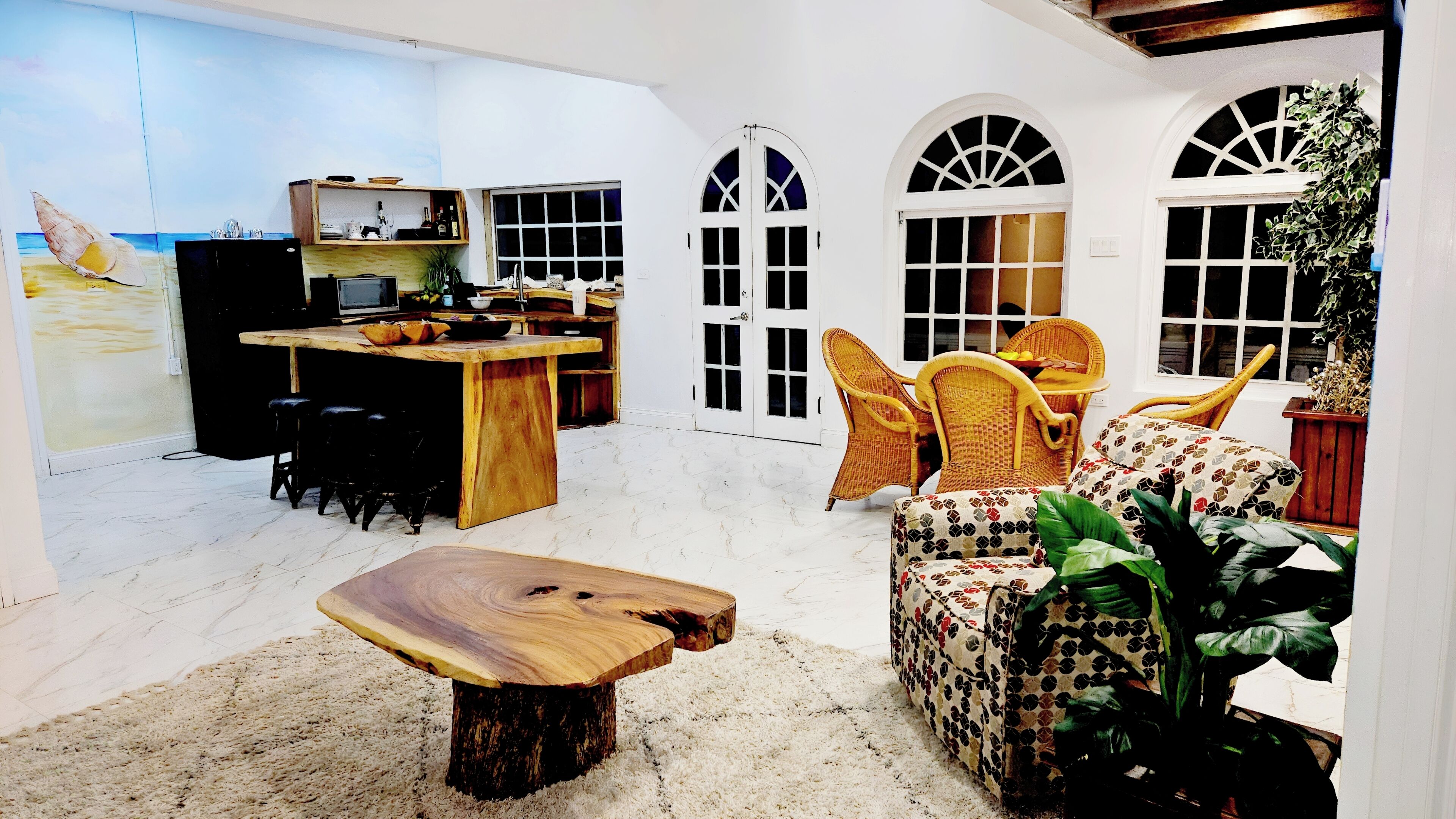 A spacious living area with a wooden coffee table, wicker chairs, a sofa, and a kitchen area with a TV and bar.