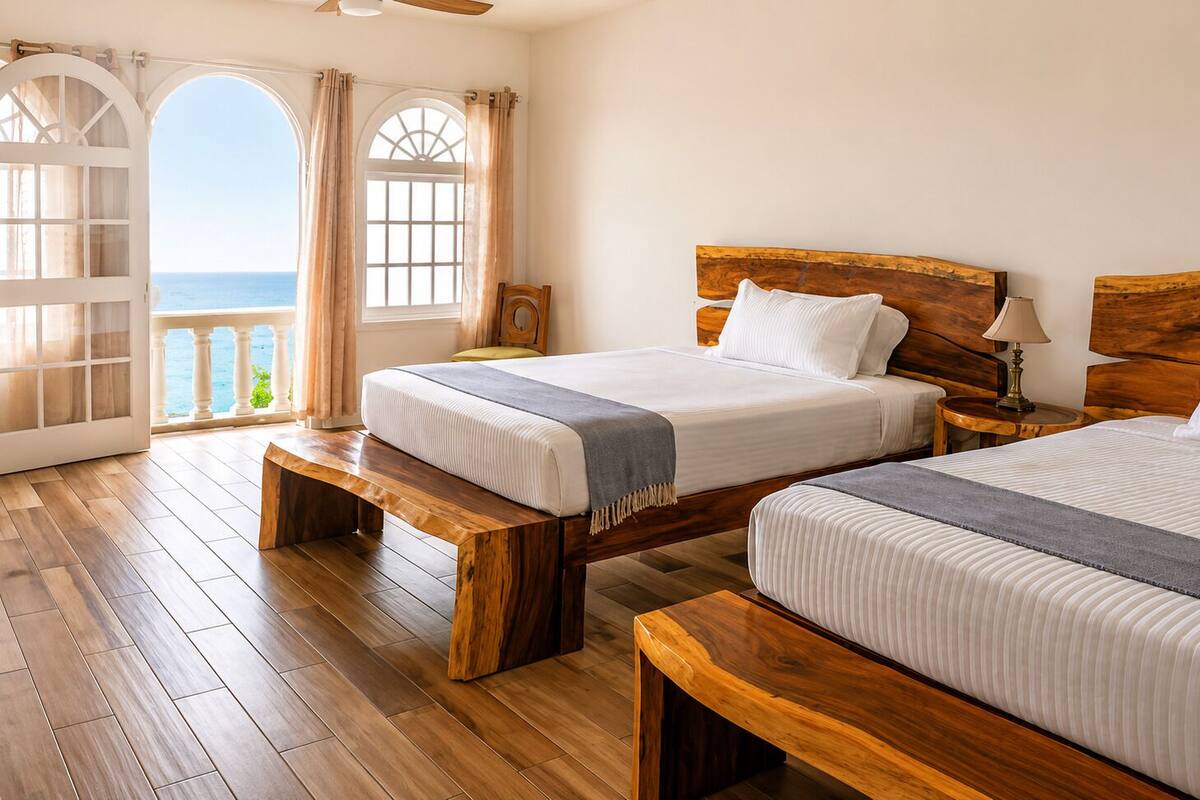 Habitación con dos camas, suelo de madera, balcón con cortinas y vista al mar.
