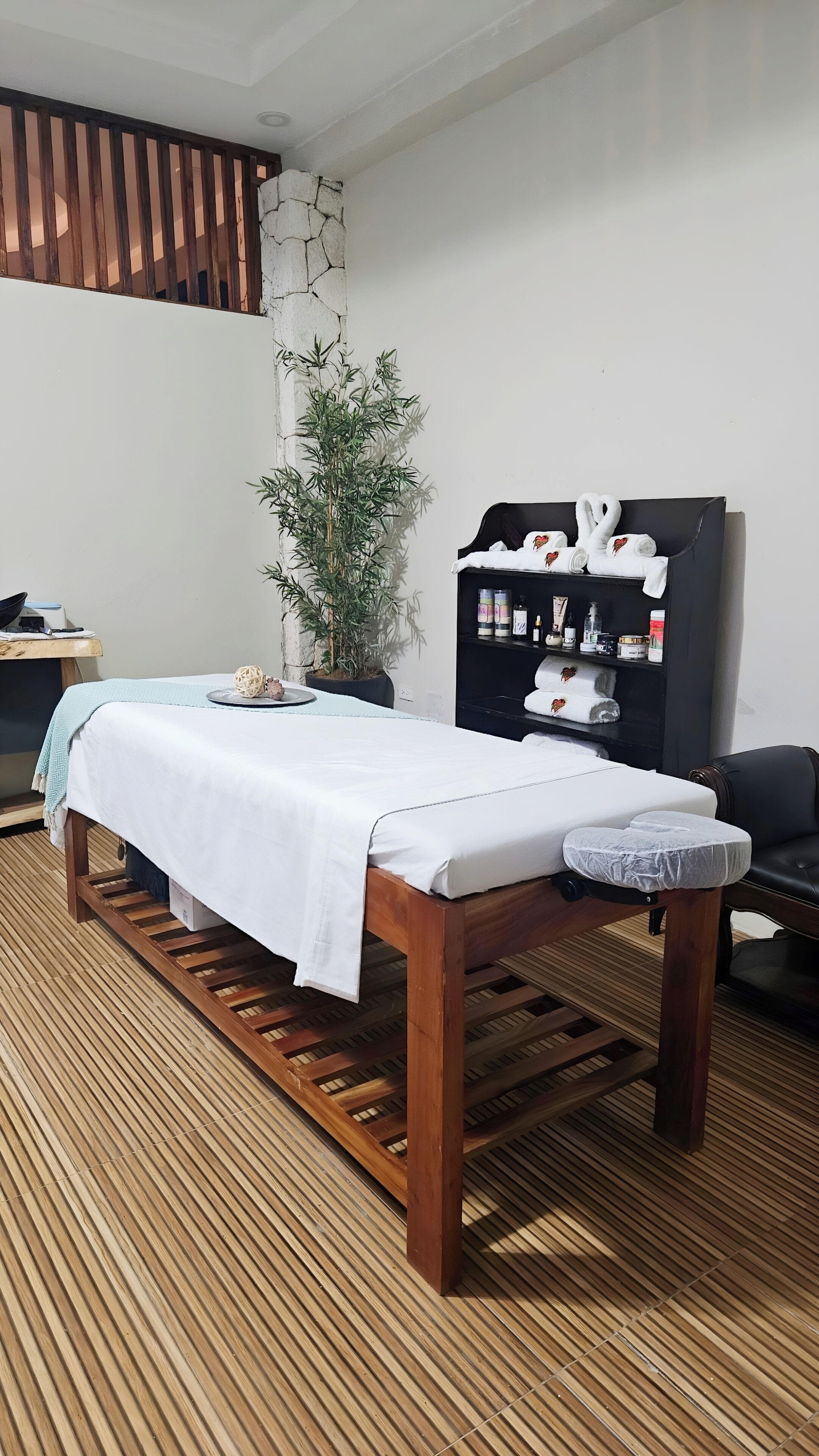 A wooden massage table with a white sheet, a bamboo plant, a shelf with wellness products, and a black leather chair.