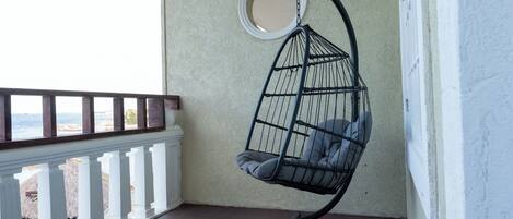A hanging chair with cushions on a balcony with a railing and a view of the sea.