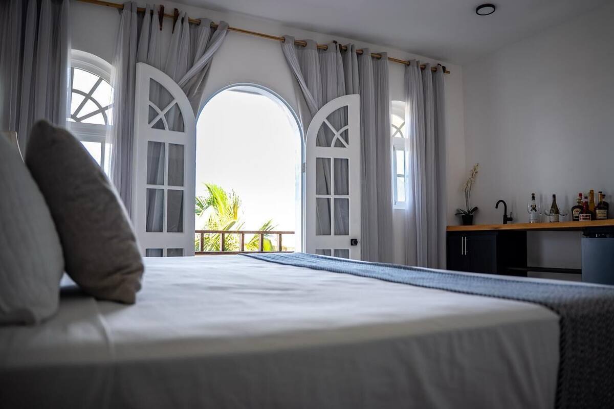 Un dormitorio con cama, cortinas, vistas por una puerta y una pequeña kitchenette.