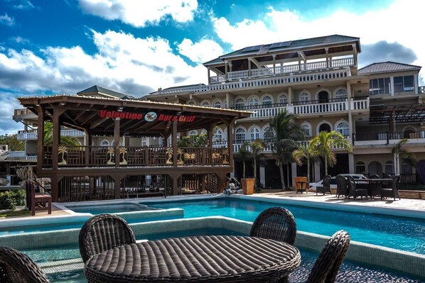 Un espace piscine avec des chaises longues, un bar appelé « Valentine's Bar & Grill », et un immeuble à plusieurs étages doté de balcons.
