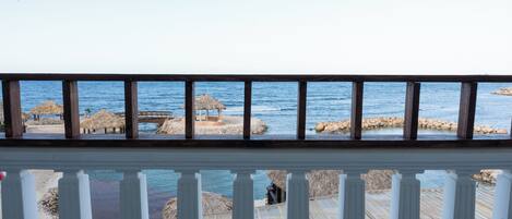 En træterrasse med gelænder, der kigger ud over en strand med stråtækte huse og en klippefyldt kystlinje.
