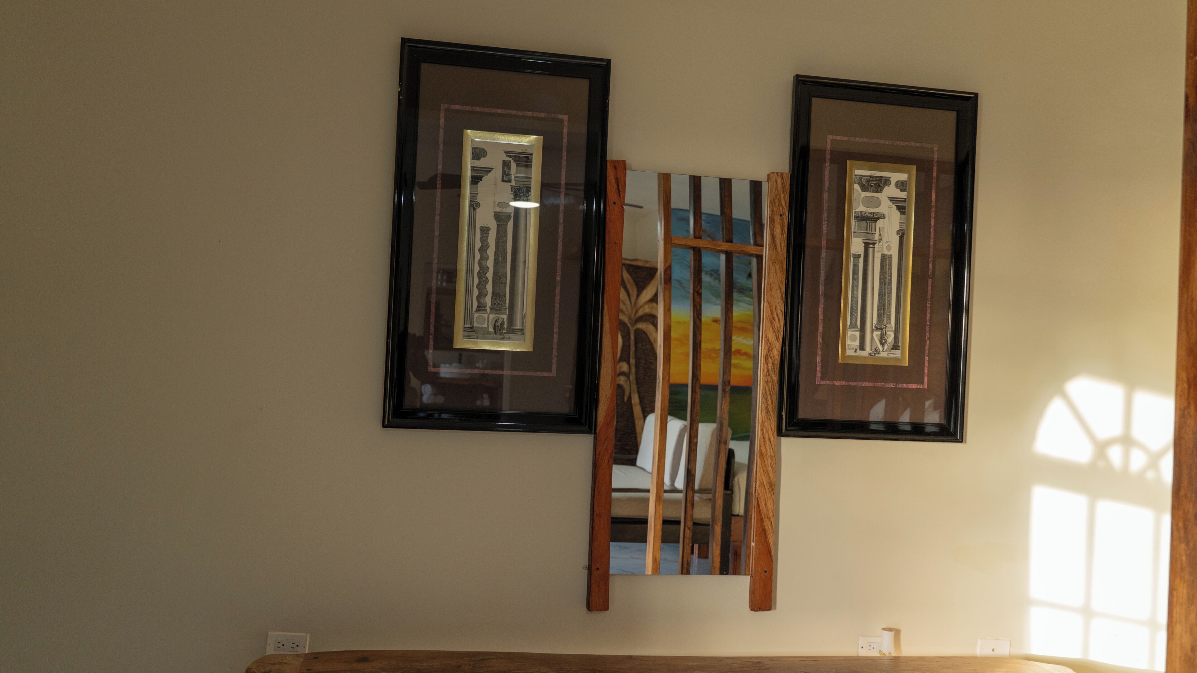 Zwei gerahmte Bilder an einer Wand, ein Holzstuhl im Vordergrund und ein Fenster, das Licht an die Wand wirft.