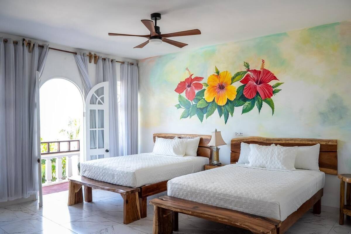 Una habitación con dos camas de madera, un ventilador de techo, un mural de flores y un balcón con cortinas.