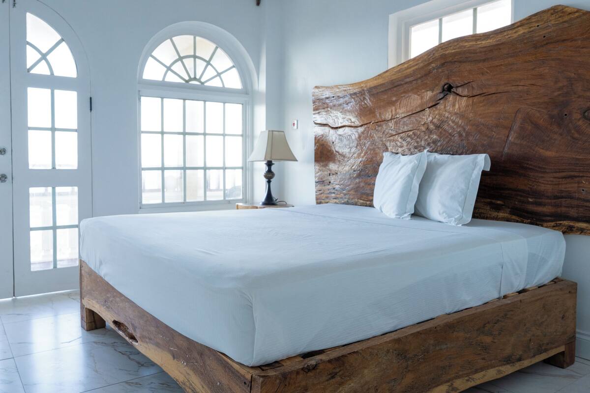 Un dormitorio con cabecera de madera, cama con ropa de cama blanca, mesita de noche con lámpara y un amplio ventanal de arco.