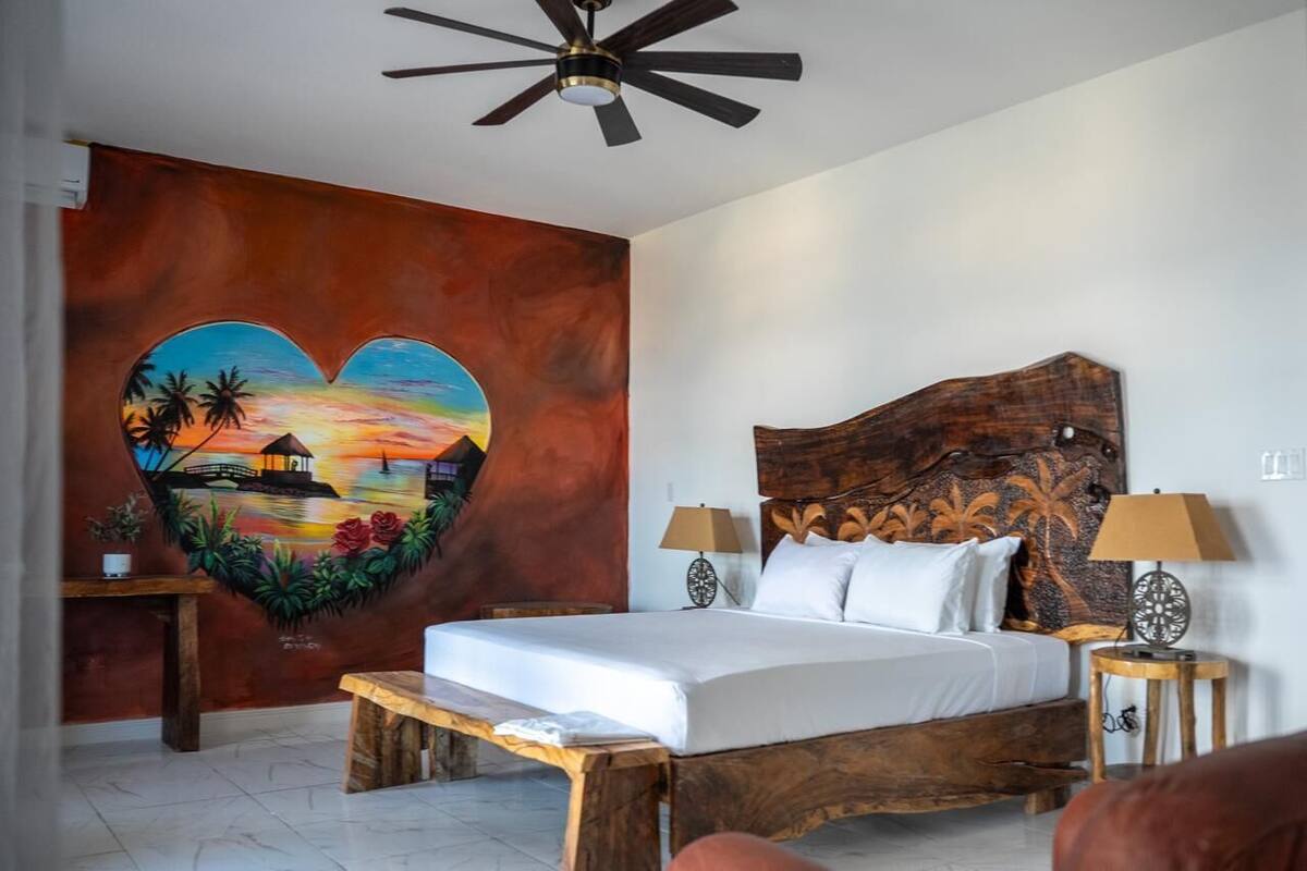 Un dormitorio con una cama, dos mesitas de noche, un ventilador de techo y un mural en la pared.