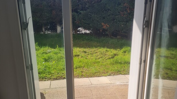 Vista a través de una puerta de cristal que muestra un área con césped, un sendero pavimentado y un edificio con techo a dos aguas al fondo.