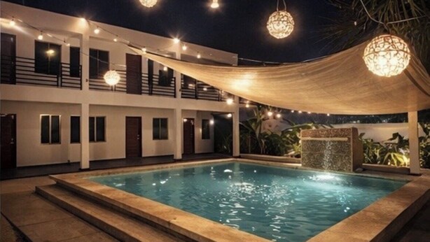 Una piscina con zona de borde de piscina, área de estar cubierta y luces colgantes.