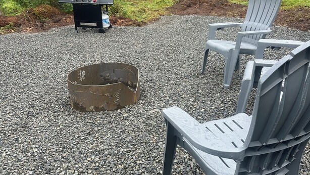Ein Kiesplatz mit Adirondack-Stühlen, einem Grill und einer Feuerstelle.
