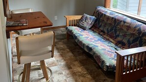 Ein kleiner, gemütlicher Raum mit einem Holztisch, einer Couch, einem Sessel und einem Fenster mit Jalousien.