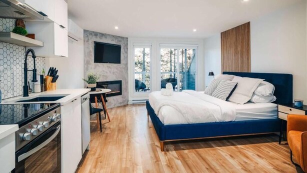 A modern bedroom with a blue bed, a small kitchen area, and a view of the outdoors. - LAO LOFT 105 by Gestion ELITE (Mont-Tremblant)