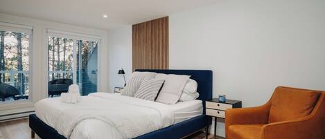 A modern bedroom with a large bed, a nightstand, a chair, and a view of trees through large windows.