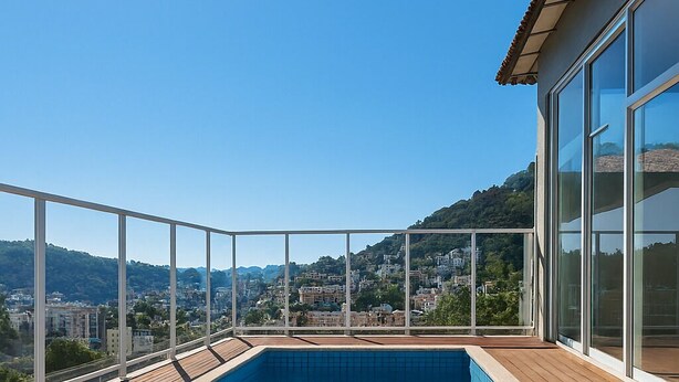 A rooftop swimming pool with a wooden deck, surrounded by glass railings, overlooking a cityscape.