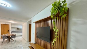 A modern living room with a flat-screen TV mounted on a wooden panel wall, a dining area with a marble floor, and a kitchen in the background.