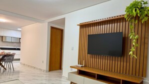 A modern living room with a flat-screen TV mounted on a wooden wall unit, a dining area with a table and chairs, and a hallway leading to a wooden door.
