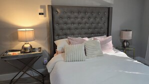 Ein Hotelzimmer mit einer gepolsterten Kopfteile, einem Nachttisch mit Lampe, einem Glastisch und einem Fenster mit Vorhängen.