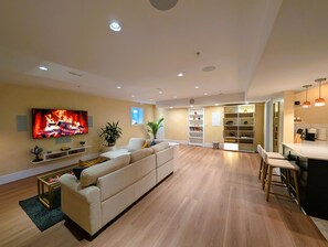 Un soggiorno moderno con una grande TV, un divano, un'area pranzo con sgabelli da bar e una cucina con luci a sospensione.