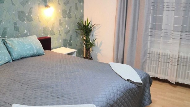 Quarto com cama, mesa de cabeceira, luminária na parede, planta em vaso e janela com cortinas.