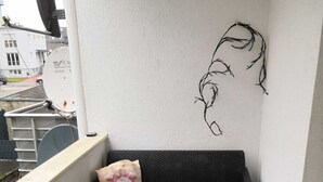 Uma varanda com um sofá de vime preto, duas almofadas, uma mesa pequena e uma obra de arte fixada na parede.