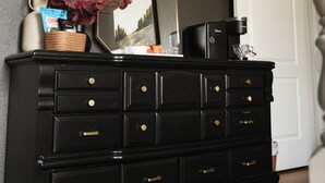 Une commode noire avec des tiroirs, une machine à café, un panier rempli de fleurs rouges et un tableau encadré accroché au mur.