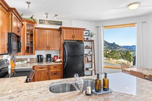 Eine Küche mit Holzschränken, einem schwarzen Kühlschrank, Granit-Arbeitsplatten, einer Spüle und Blick auf Berge durch ein großes Fenster.