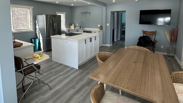 Una cocina moderna con armarios blancos, una isla central y un comedor con mesa de madera y sillas de mimbre.